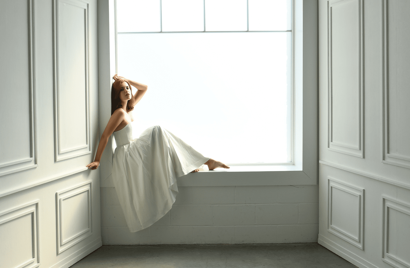 A redheaded senior girl in a white gown sits in the window sill during her senior photo session at Kliks Photography.