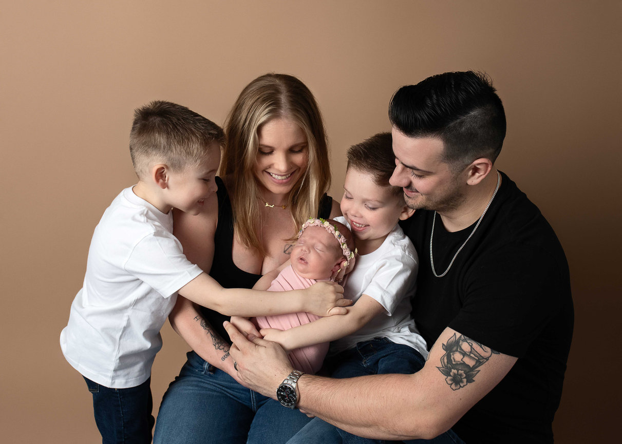 Smiling family of five cuddles newborn baby girl swaddled in pink with floral headband – newborn family photography in Dundee, Illinois