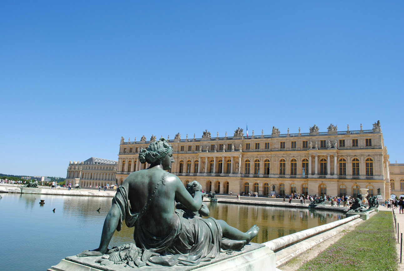 Statue by a pond facing the Palace of Versailles, with a clear blue sky.