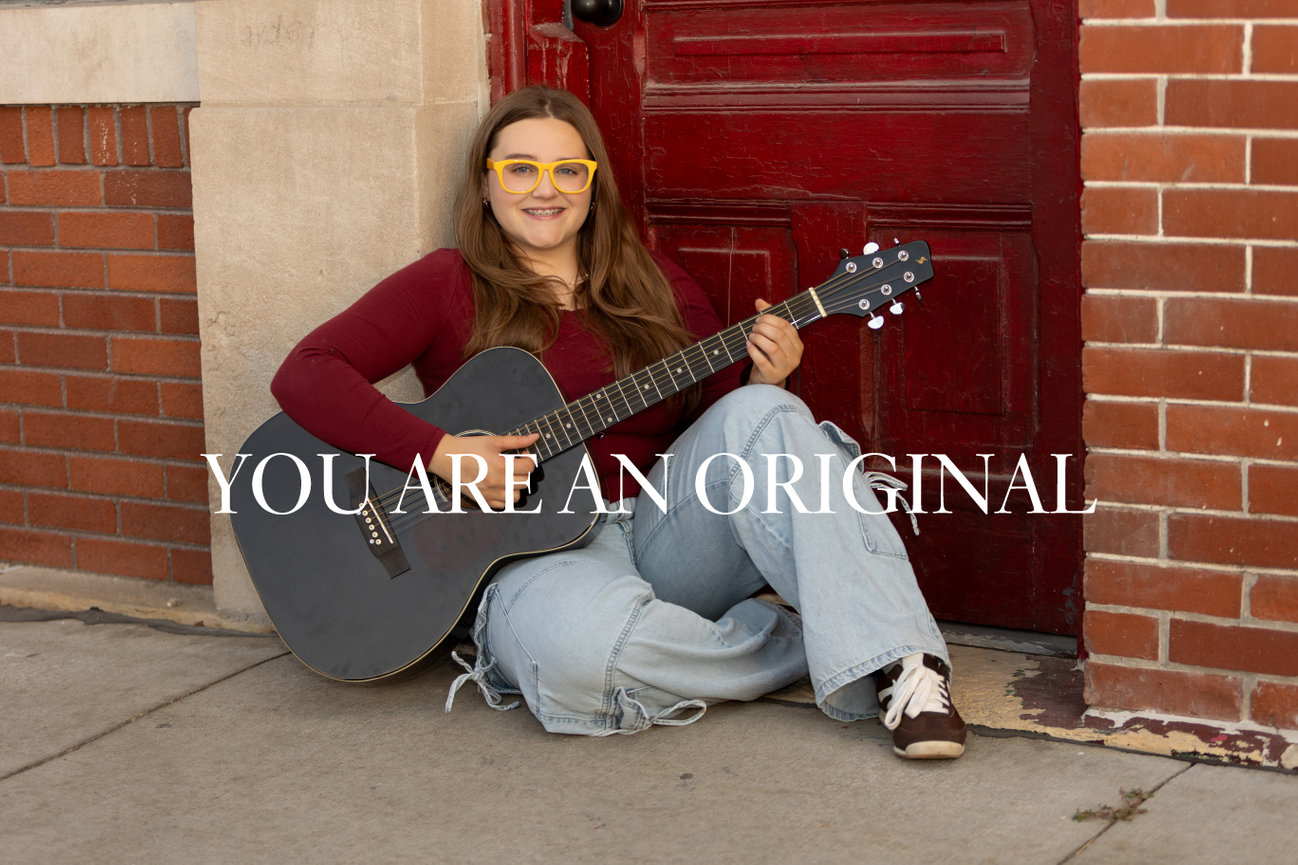 A person with glasses plays an acoustic guitar while sitting by a red door. Text reads YOU ARE AN ORIGINAL.