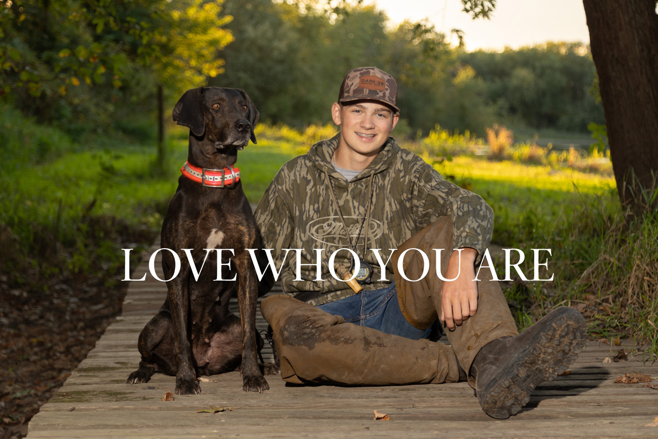 Teen in camo outfit sits on a wooden path with a dog, surrounded by trees and grass. Text reads LOVE WHO YOU ARE.