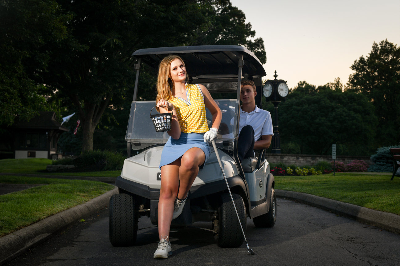 A young woman poses confidently beside a golf cart, holding clubs, while a man sits in the cart.