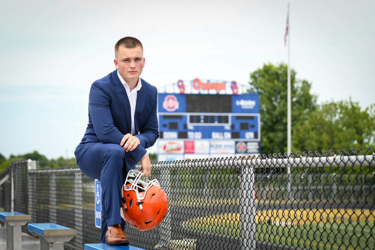 A young man in a blue suit sits on a bleacher holding an orange football helmet, with a scoreboard in the background.