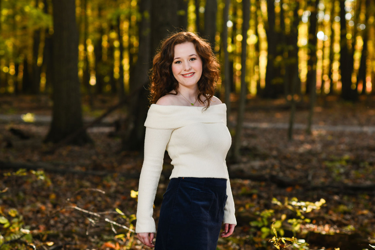 Senior girl standing in the middle of a wooded area during fall, wearing a mini skirt and off-the-shoulder sweater