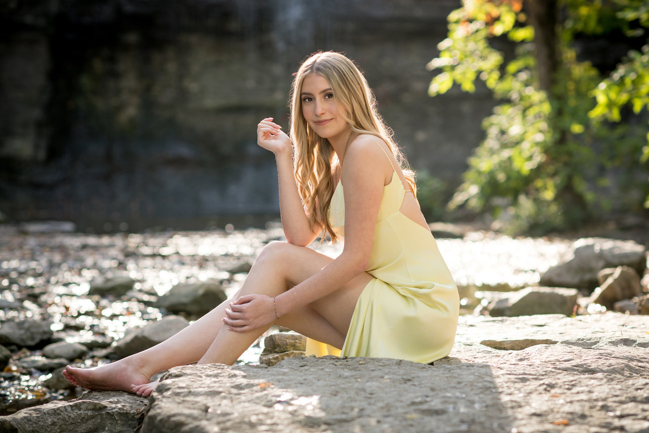 Teen girl in a yellow dress sitting on a rock by a stream, surrounded by greenery and waterfalls for senior pictures in Columbus Ohio.