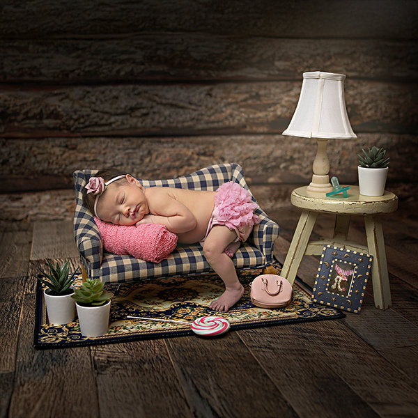 Baby sleeping on a small couch with a lamp, plants, and toys on a wooden floor background.