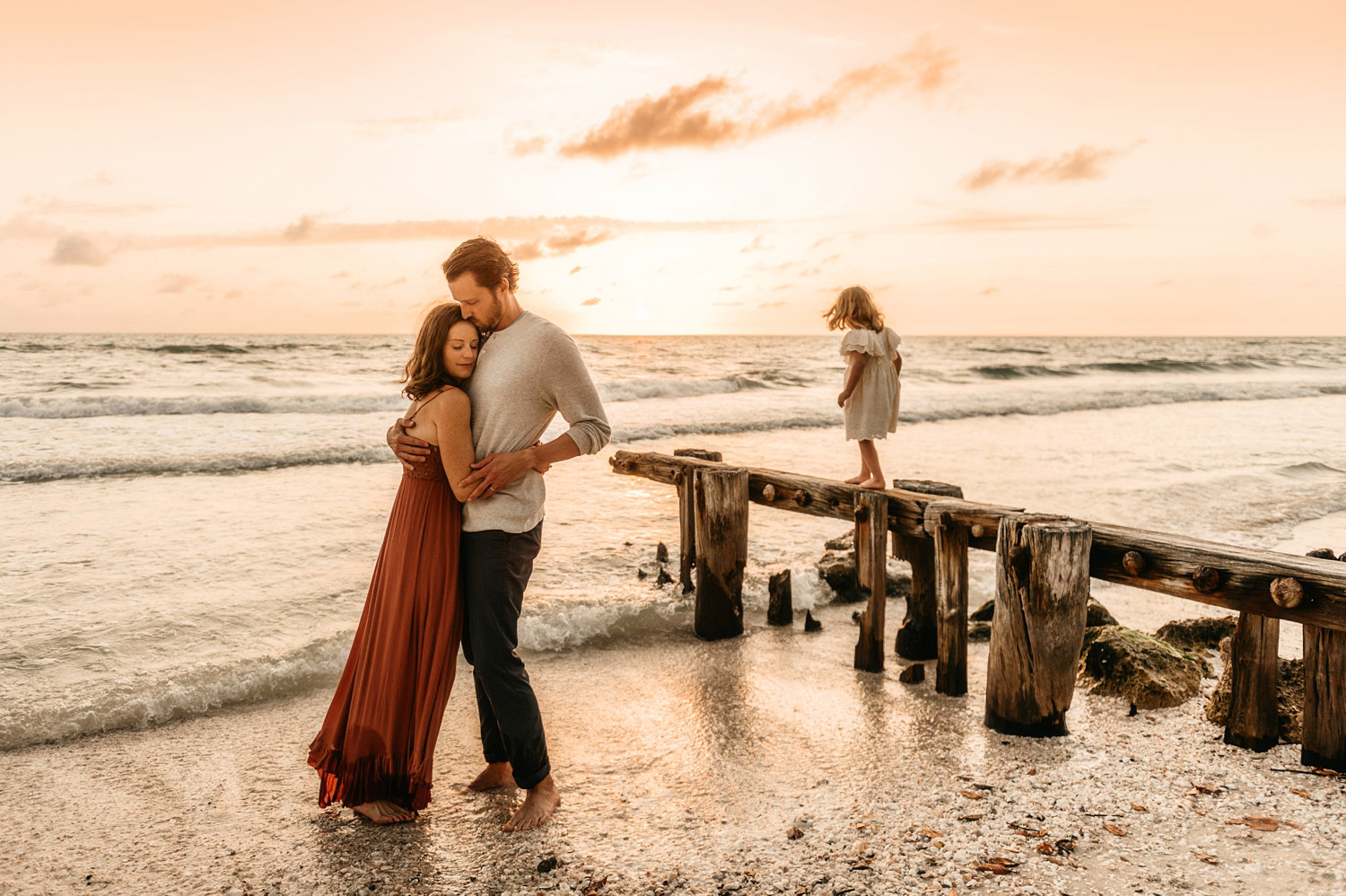 mom and dad hugging at the beach while little girl plays in background, Rya Duncklee Photos